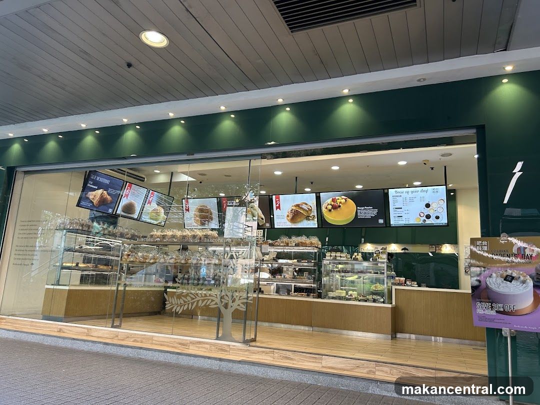 A wide shot of a bakery's storefront, showcasing its interior through large glass windows. Display cases filled with pastries and cakes are visible, along with digital menu boards above. The exterior features a dark green facade with subtle lighting. A promotional sign for Parents' Day is placed to the right.