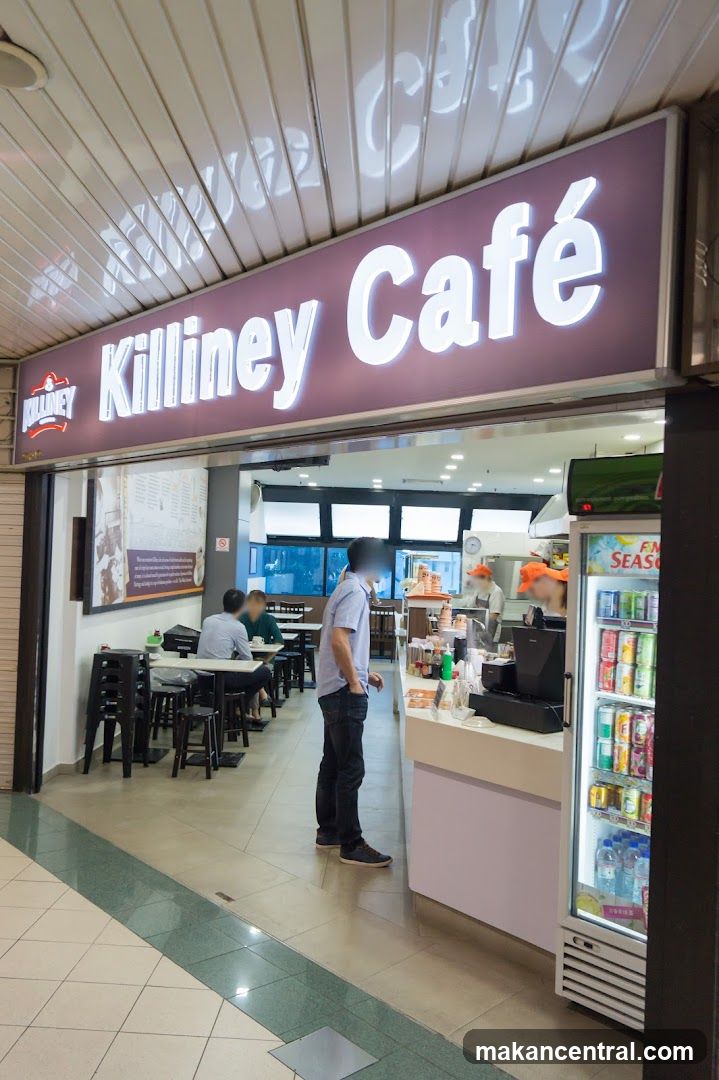 The exterior of a Killiney Cafe with a prominent illuminated sign. Inside, tables and chairs are set up for dining, and staff can be seen behind a counter. A refrigerator stocked with drinks is visible to the right.