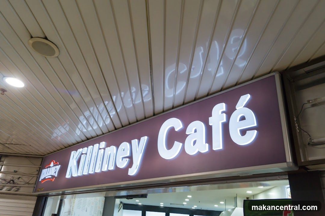 A lit sign with the words "Killiney Café" in white, sans-serif font against a dark purple background. The sign is mounted above an entrance. A smaller logo for "Killiney" is visible on the left side of the sign.