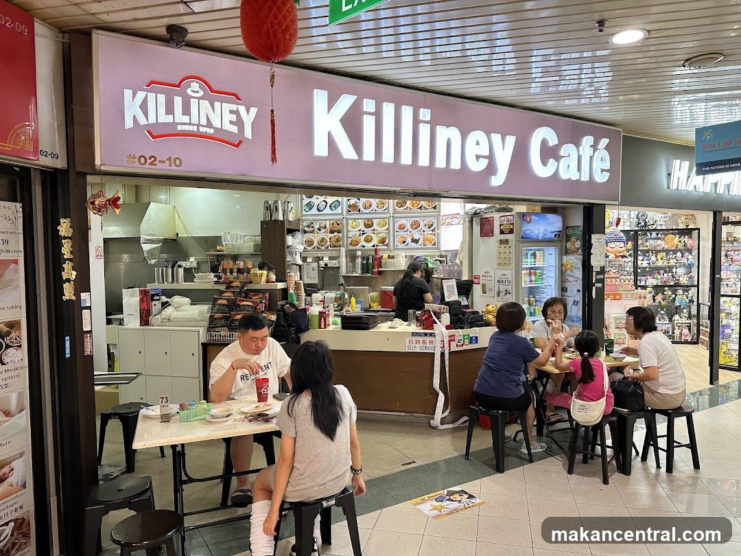The exterior of a Killiney Cafe with a large illuminated sign. People are seated at tables inside and outside the cafe. The cafe is located in a mall with other shops visible.