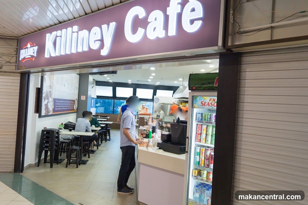 The exterior of Killiney Cafe shows a wide storefront with a large illuminated sign above the entrance. Inside, tables and chairs are visible, with customers seated. A counter area with a refrigerator stocked with drinks is also present.