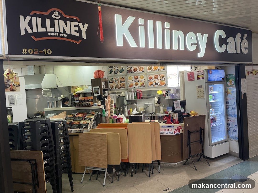 The exterior of a Killiney Cafe with a large illuminated sign above the entrance. The cafe has a display of food pictures on the wall behind the counter. Stacks of chairs and tables are visible in front of the cafe. A refrigerator stocked with drinks is on the right side of the entrance.