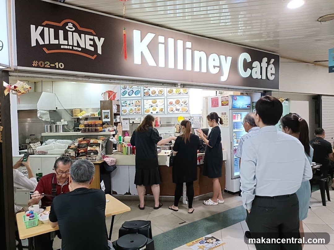 A busy Killiney Cafe with customers ordering and dining. The cafe has a prominent sign with the cafe's name and logo. Behind the counter, food is displayed on a menu board. People are seen queuing and eating at tables.