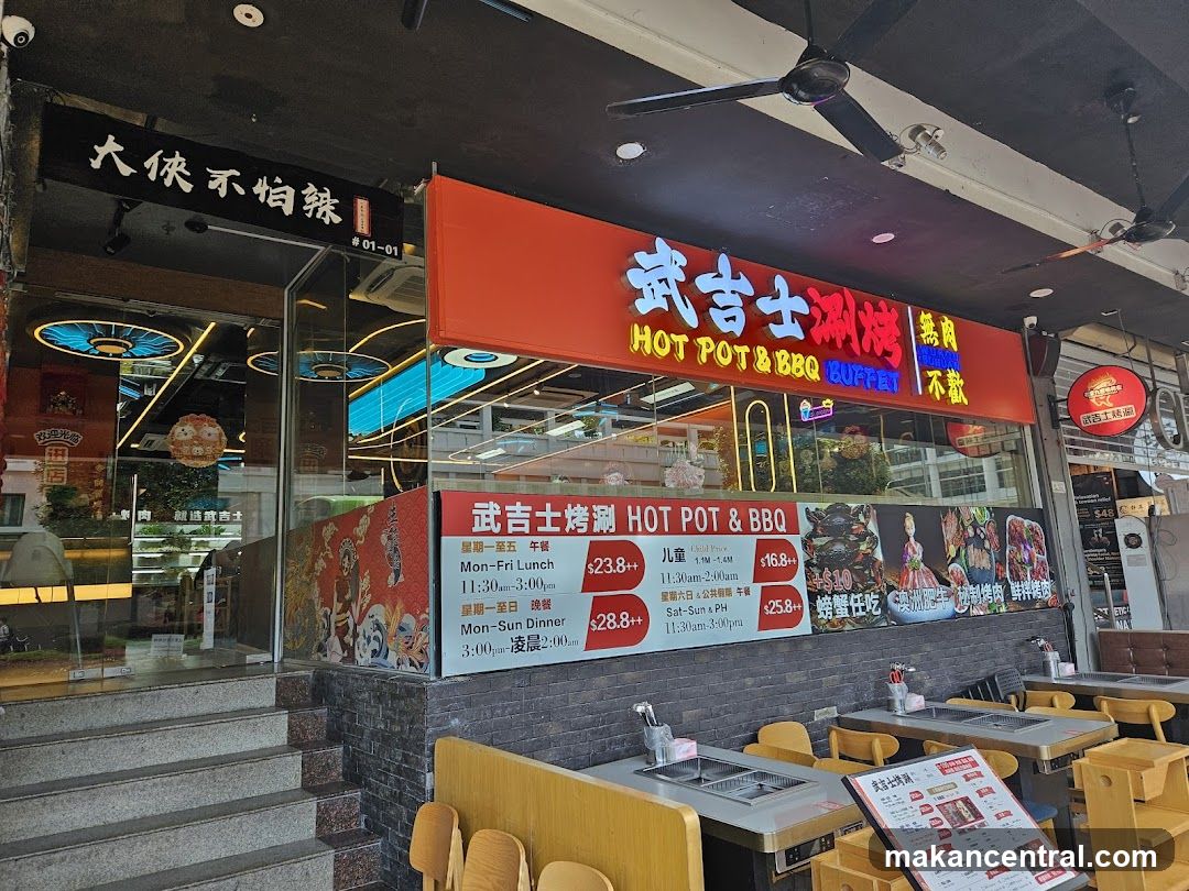 The exterior of a restaurant with a prominent red awning displaying "HOT POT & BBQ BUFFET" in large neon letters. Below the awning, a menu board lists prices for lunch and dinner, with options for weekdays and weekends. The entrance is visible with stairs leading up to it. Tables with built-in grills are set up outside the restaurant.