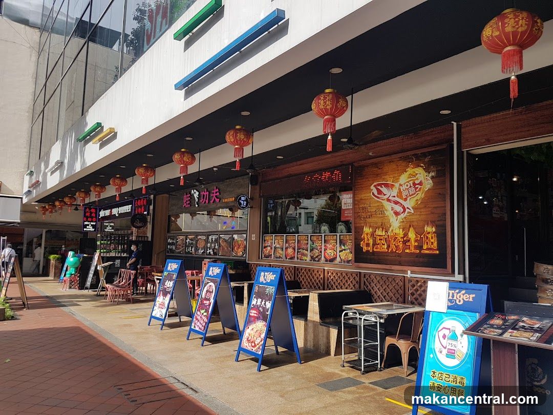 A row of restaurants with outdoor seating and signage. Red lanterns hang overhead. Several A-frame signs display menus and promotions. One sign features a Tiger beer logo. The facade includes large windows and wooden paneling with graphics.