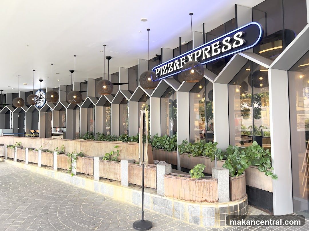The exterior of a Pizza Express restaurant with a prominent neon sign above the entrance. The storefront features large glass windows and a modern architectural design with geometric patterns. Planters with lush green plants line the walkway in front of the restaurant.