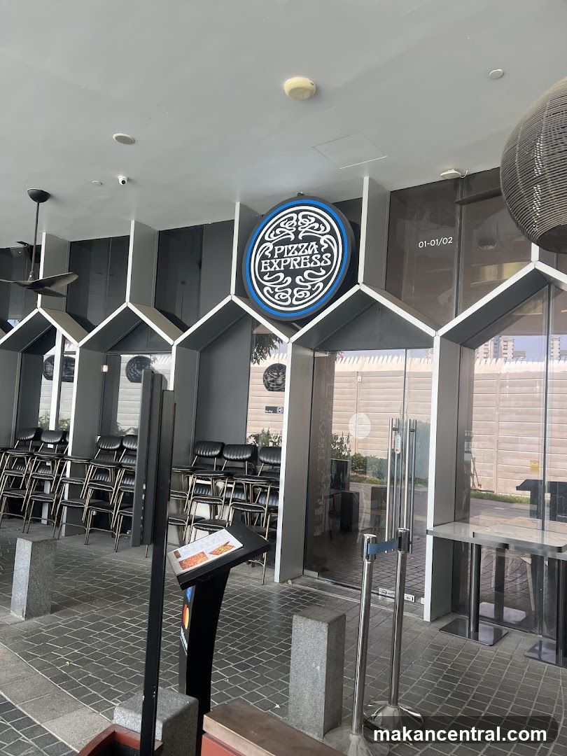 The exterior of a Pizza Express restaurant. A circular sign with the restaurant's logo is visible above the entrance. Stacked chairs are visible through the glass doors.