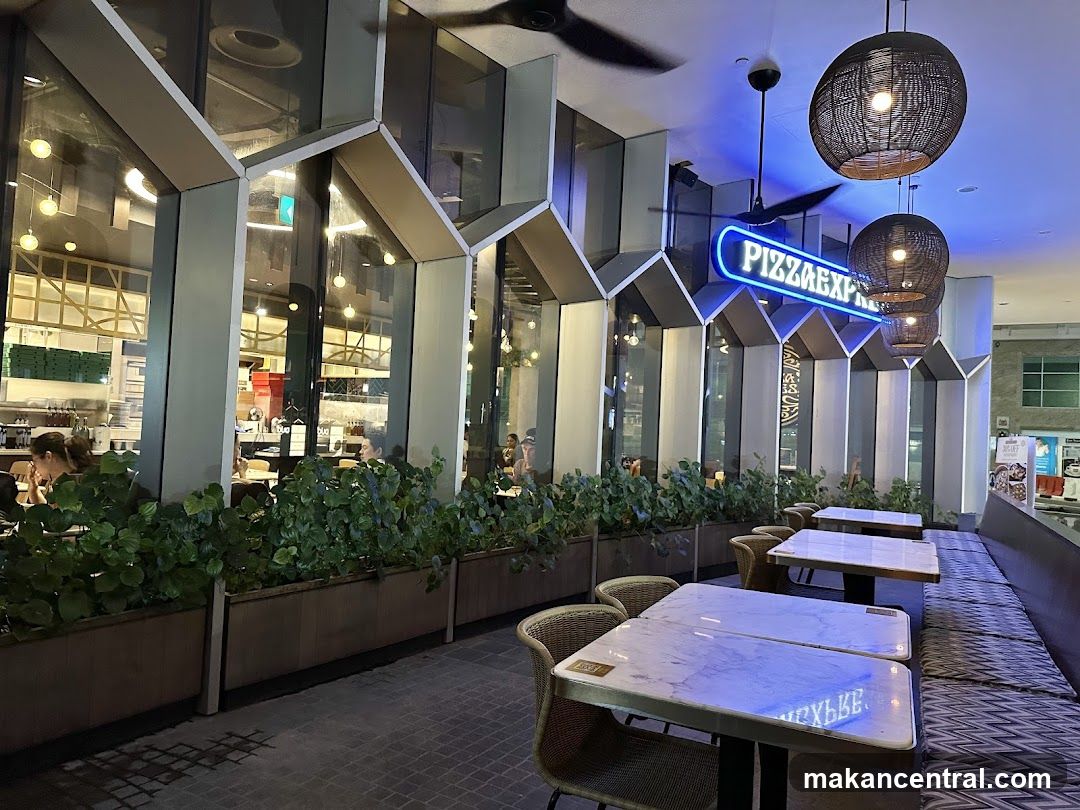 The exterior of a Pizza Express restaurant at night. The restaurant has large windows and a neon sign that says "PIZZAEXPRESS". Inside, tables and chairs are visible, as well as people dining. There are also hanging lamps and a ceiling fan.