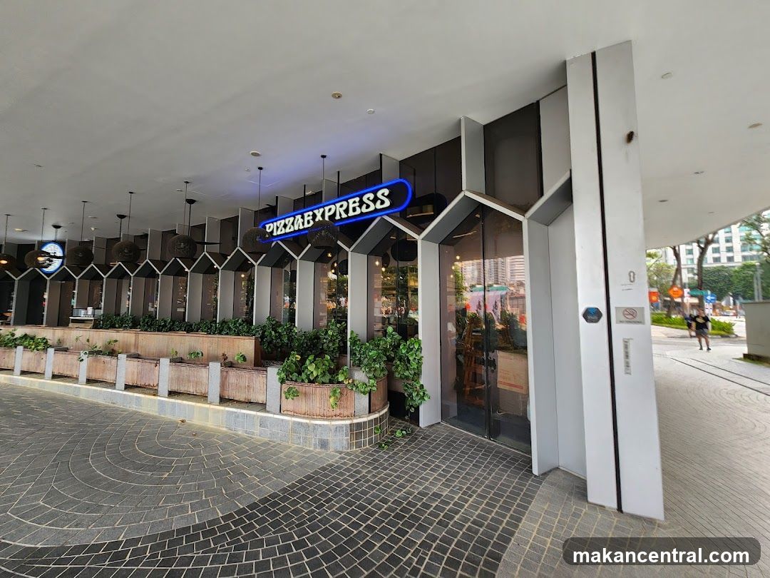 The exterior of a Pizza Express restaurant with a prominent neon sign above the entrance. The storefront features large glass windows and is lined with planters containing greenery. The walkway in front is paved with grey tiles arranged in a circular pattern.