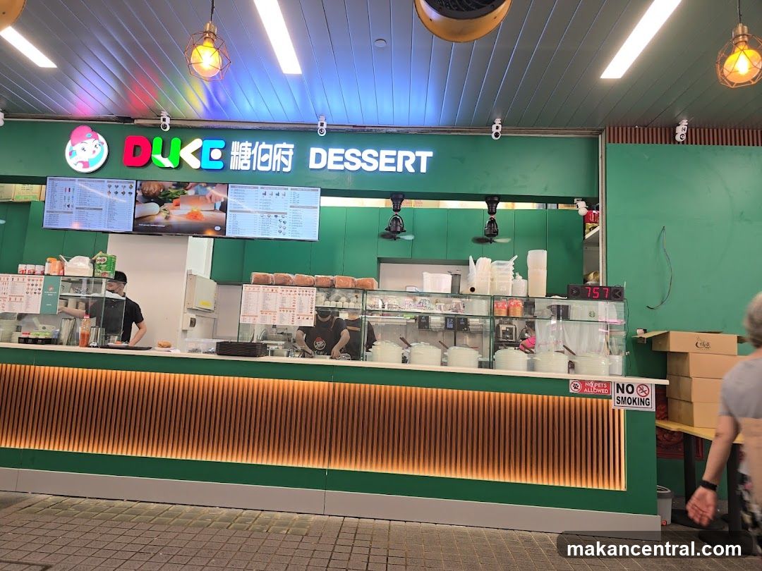 A green storefront with the DUKE DESSERT logo and signage. Digital menus are displayed above the counter. The counter area shows food preparation and service.
