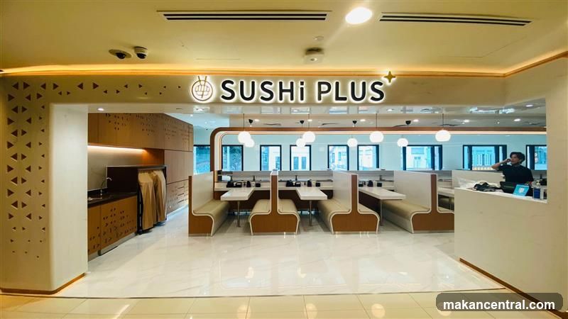 The image shows the interior entrance of a restaurant called Sushi Plus. The restaurant features booth seating, tables, and a counter area. The signage "SUSHI PLUS" is prominently displayed above the entrance. The decor is modern and clean, with warm lighting and light-colored seating. A person is visible behind the counter on the right side of the image.
