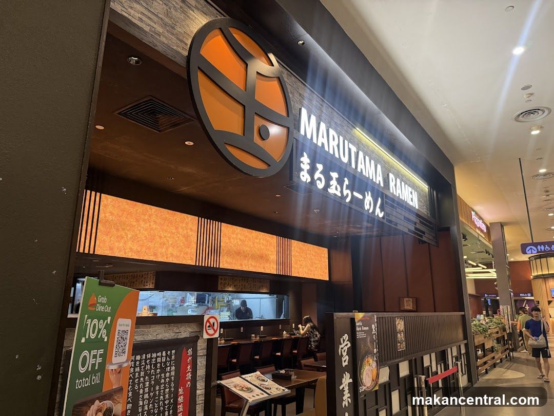 The exterior of a Marutama Ramen restaurant. The shop front features a large circular logo and a lit sign with the restaurant name in English and Japanese. The interior is visible through the open entrance, showing tables and a kitchen area. A promotional flyer for Grab Dine Out offering 10% off is displayed near the entrance.