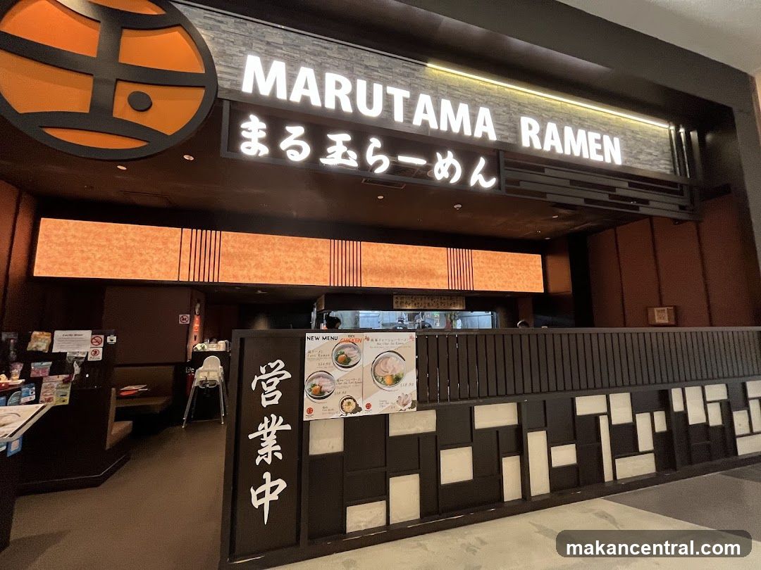 The exterior of a Marutama Ramen restaurant. The prominent signage displays "MARUTAMA RAMEN" in large white letters, with Japanese characters below. A circular logo with orange and black elements is visible to the left of the main sign. The entrance area features warm lighting and a dark wooden facade. A menu board is partially visible to the right, displaying images of ramen bowls and prices.