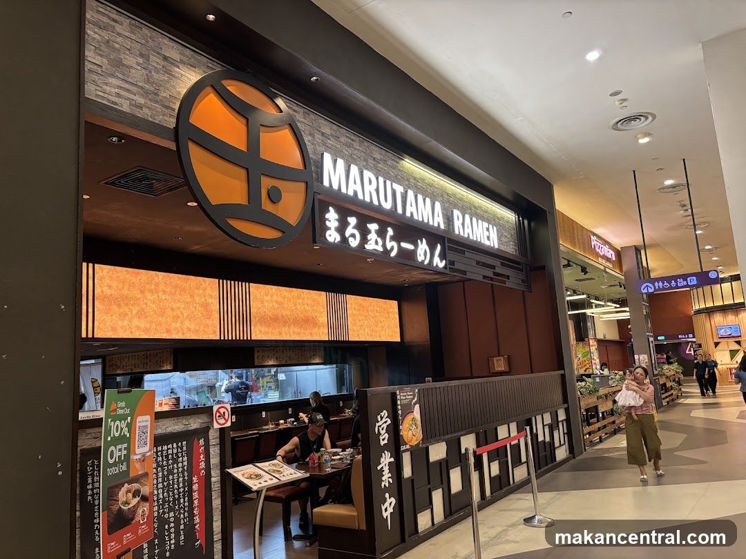 The exterior of a Marutama Ramen restaurant with its prominent signage. The restaurant has a modern facade with dark wood accents and illuminated panels. Inside, diners can be seen seated at tables, and the kitchen area is visible. A promotional flyer for a 10% discount is displayed near the entrance.
