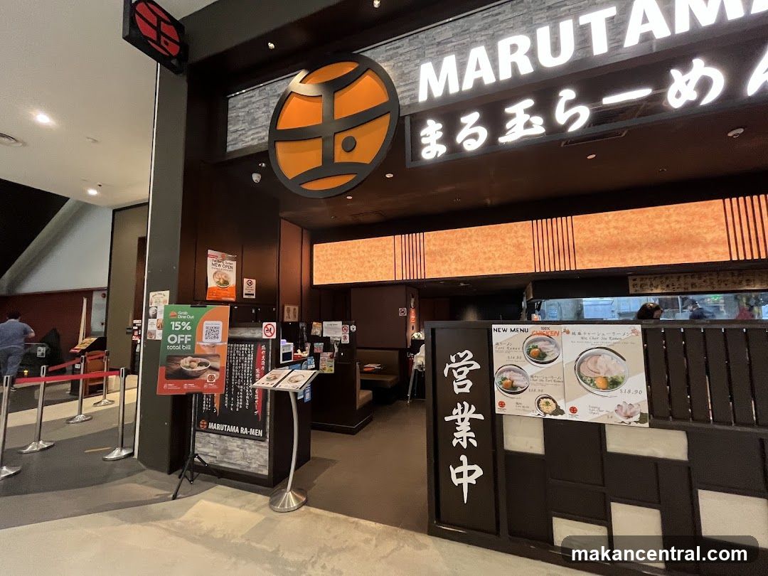 The exterior of a Marutama Ra-Men restaurant with its prominent signage. A promotional sign offering 15% off the total bill is displayed near the entrance.