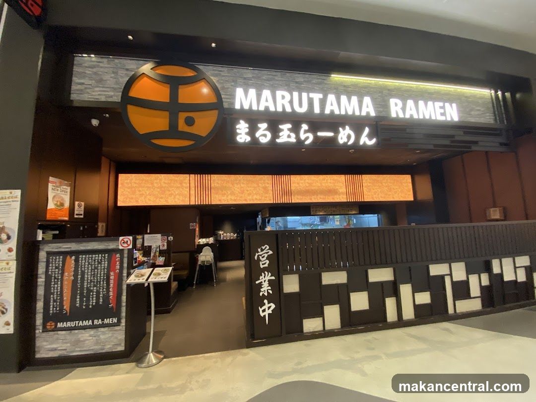 The exterior of a Marutama Ramen restaurant. A large circular logo with orange and black accents is visible above the entrance. The restaurant name "MARUTAMA RAMEN" is displayed in white illuminated lettering. Below the main sign, Japanese characters are also visible. To the left of the entrance, a menu board displays text and images. The entrance itself is open, revealing seating and a counter area inside.