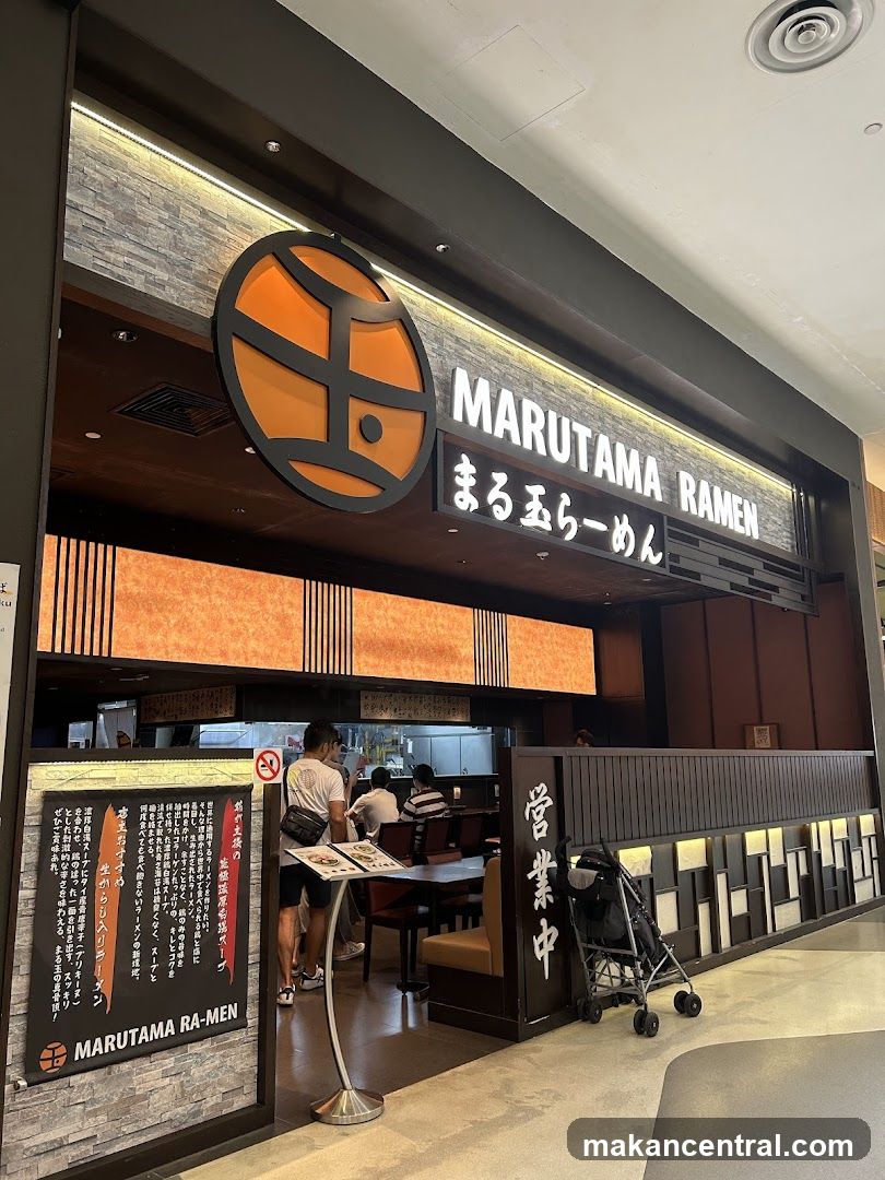 The exterior of a Marutama Ramen restaurant. A large circular logo with orange and black elements is visible above the entrance, along with the text "MARUTAMA RAMEN" in white lettering. Below the main sign, Japanese characters are also displayed. A menu board with Japanese text and the restaurant's name is positioned to the left of the entrance. The interior of the restaurant, including seating and kitchen staff, is visible through the open doorway. A stroller is parked to the right of the entrance.