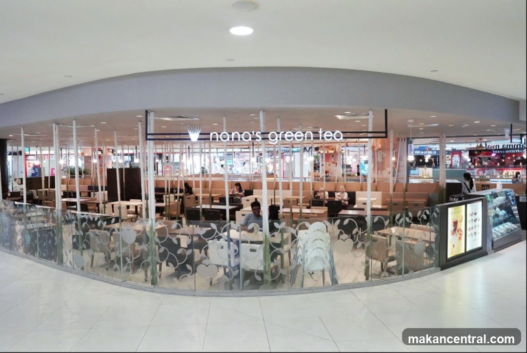 A wide shot of the exterior of a Nana's Green Tea shop located in a mall. The shop has a modern design with a glass facade and interior seating visible. The shop's name is displayed prominently above the entrance. Inside, tables and chairs are arranged, and some customers are seated. A display counter with food items is visible to the right.