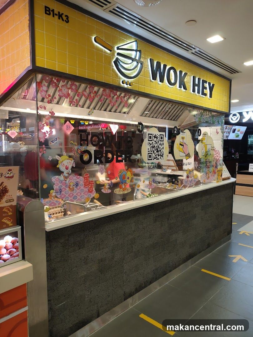 The exterior of a Wok Hey food stall with a yellow tiled facade and a prominent "WOK HEY" logo. The counter area has glass windows with decorative stickers and instructions on how to order. A QR code is visible for scanning.