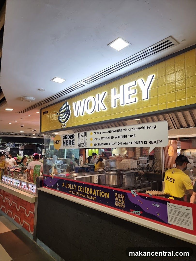 The exterior of a Wok Hey food stall with a bright yellow tiled facade and prominent white lettering spelling out the brand name. Below the main sign, a white speech bubble graphic provides ordering instructions and information about checking wait times and receiving notifications. A colorful banner with the text "A JOLLY CELEBRATION OF EXCEPTIONAL TALENT" is displayed across the front counter. Staff members in yellow shirts with "HEY" printed on them are visible behind the counter.