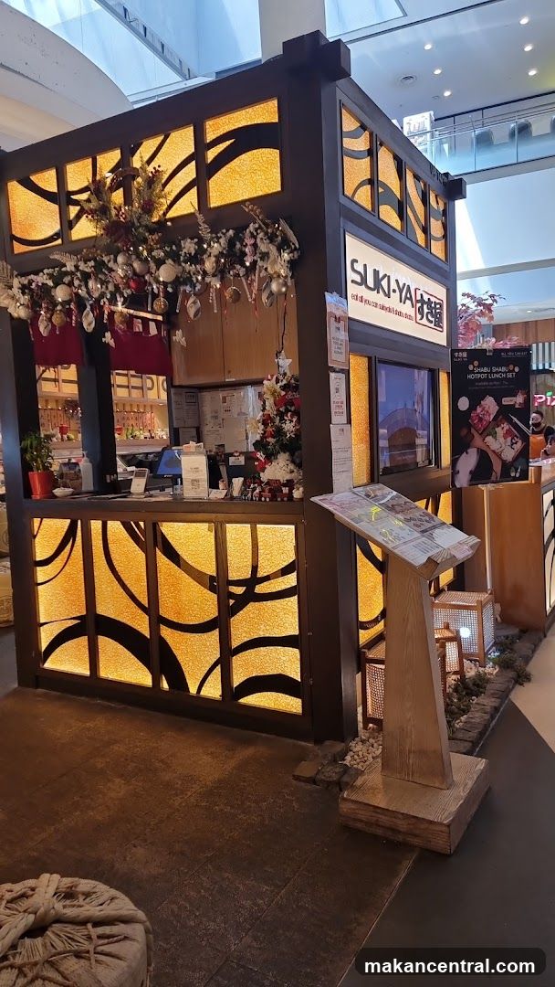 The exterior of a Suki-Ya restaurant stall, decorated with festive Christmas garlands and lights. The stall has a dark wood facade with illuminated panels featuring a wavy pattern. A prominent sign above the counter reads "SUKI-YA" with Japanese characters. A menu stand is placed in front of the stall.