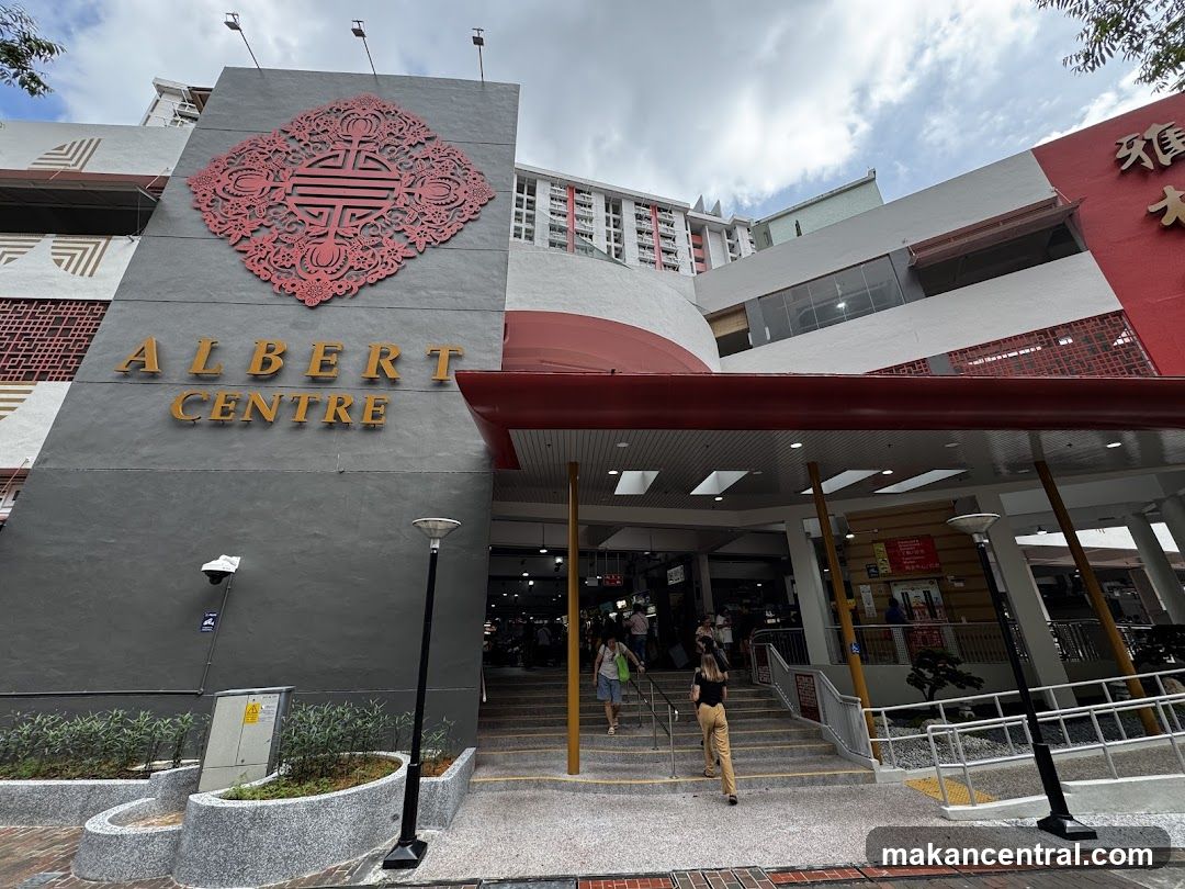The exterior of the Albert Centre building, featuring prominent signage with the name "ALBERT CENTRE" in gold lettering against a grey facade. A large, ornate red decorative element is positioned above the signage. The entrance to the centre is visible with a set of stairs leading up to it, and several people are seen entering and exiting. The building has a modern architectural style with white and red accents.