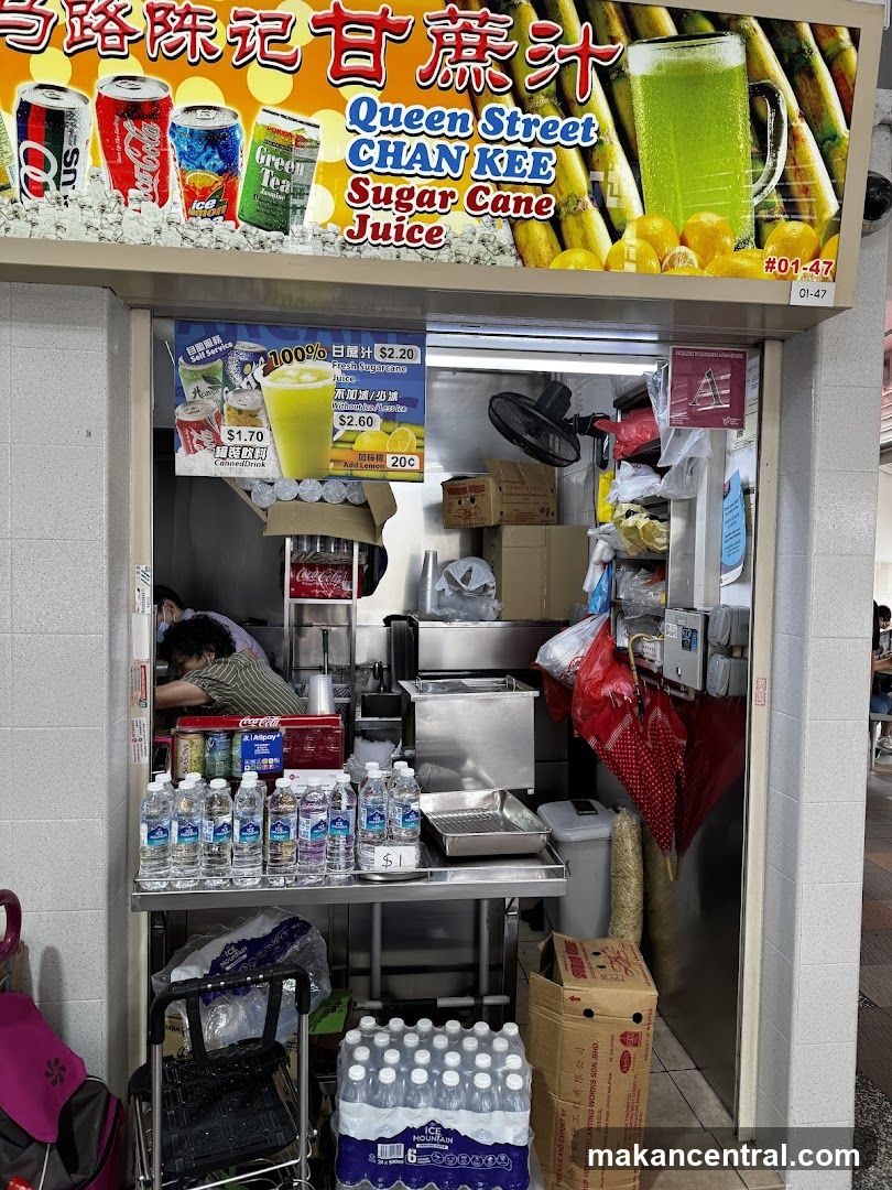 A stall selling sugar cane juice and canned drinks. The stall has a large sign with the business name and images of sugar cane and drinks. Below the sign is a menu board with prices for fresh sugarcane juice and canned drinks. Bottles of water and other drinks are displayed on shelves.