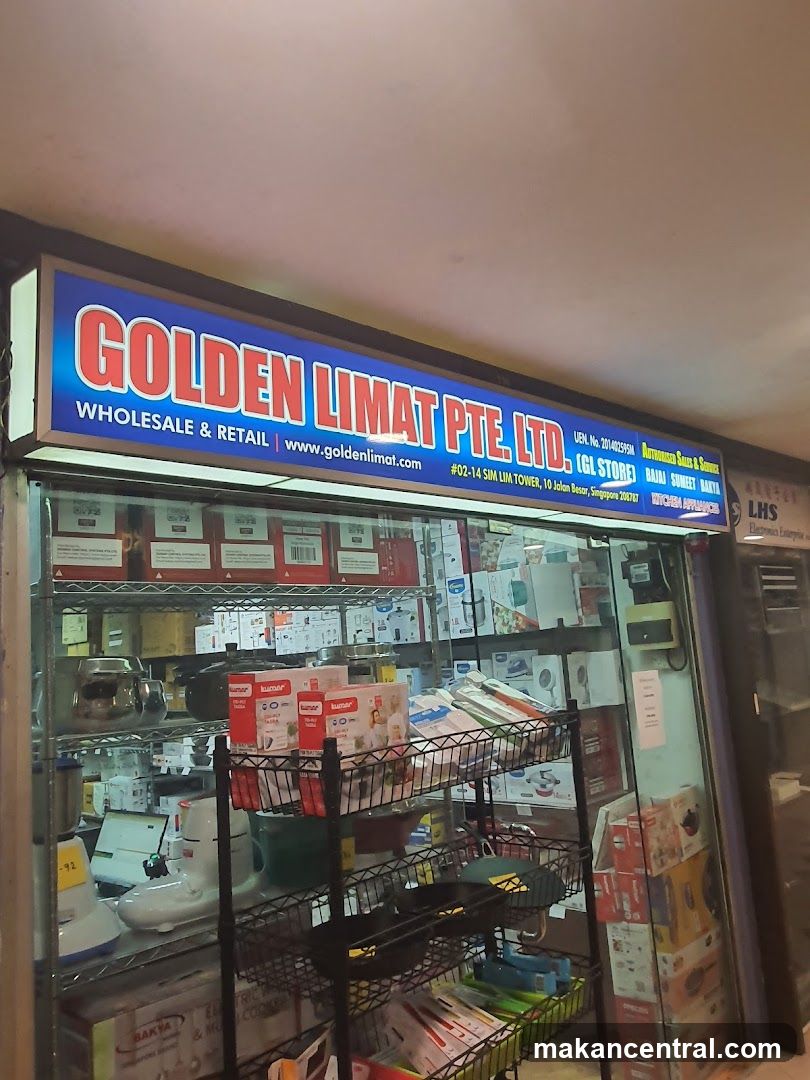 The exterior of a store with a large illuminated sign that reads "GOLDEN LIMAT PTE LTD, WHOLESALE & RETAIL" and a website address. Below the sign, the store displays various kitchen appliances and cookware on shelves and racks. The store is located at #02-14 Sim Lim Tower, 10 Jalan Besar, Singapore 206767.