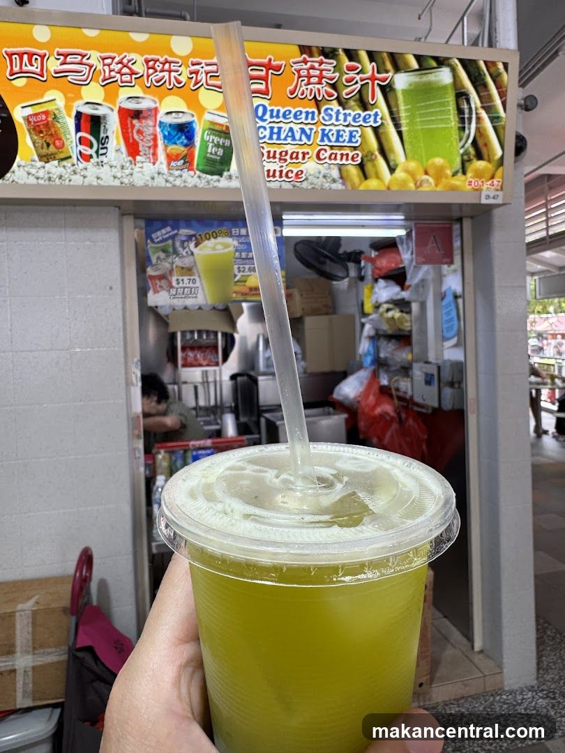 A hand holds a plastic cup filled with green sugar cane juice. Behind the cup is the storefront of Queen Street Chan Kee Sugar Cane Juice. A large sign above the stall displays the business name in Chinese and English, along with images of canned drinks and sugar cane. Below the main sign, a smaller menu board shows prices for drinks. The stall interior is visible, with a person working behind the counter.