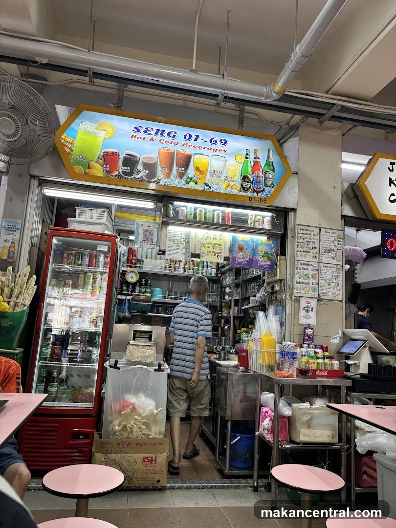 A street-level view of a beverage stall with a prominent sign displaying "SENG 01-69 Hot & Cold Beverages" and images of various drinks. A man is seen standing inside the stall, facing away from the camera. To the left, a red refrigerator is stocked with drinks. The stall appears to be part of a larger market or food court, with tables and chairs visible in the foreground.