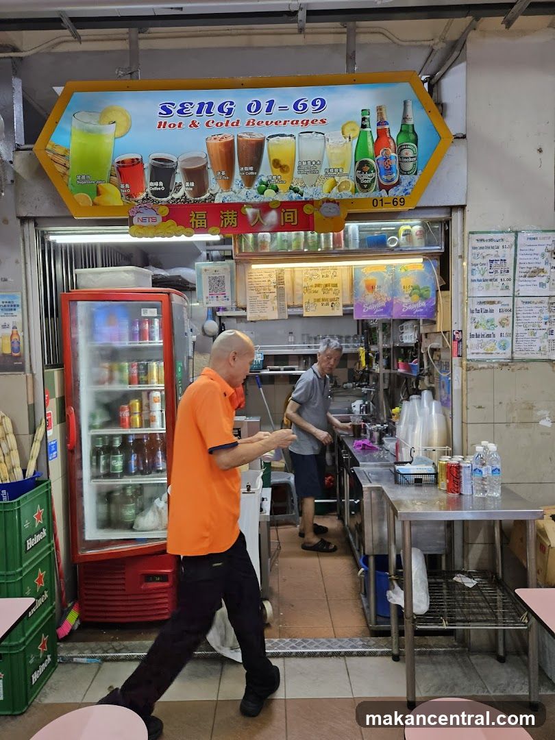 A hexagonal sign displays various hot and cold beverages, including juices, coffees, and beers, with the business name "Seng 01-69 Hot & Cold Beverages" prominently featured. Below the sign, a stall interior is visible with a red refrigerator stocked with drinks, a counter where two men are working, and various beverages and supplies.