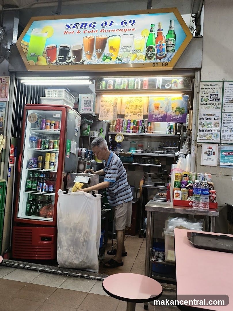 A street-level view of a beverage stall with a prominent sign displaying "SENG 01-69 Hot & Cold Beverages" and images of various drinks. A man is seen working behind the counter, and a red refrigerator is filled with drinks. Shelves are stocked with canned beverages and other items. Tables and chairs are visible in the foreground.