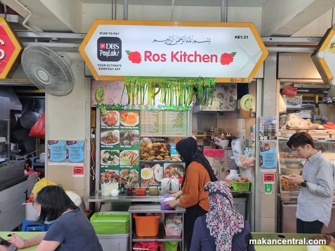 A food stall with a prominent sign that reads "Ros Kitchen" and displays a DBS PayLah! QR code for payment. The stall appears to be in a hawker center or food court, with a menu board featuring various dishes and prices. Several customers are visible, some ordering and others seated at tables. The stall is decorated with green streamers and floral motifs.