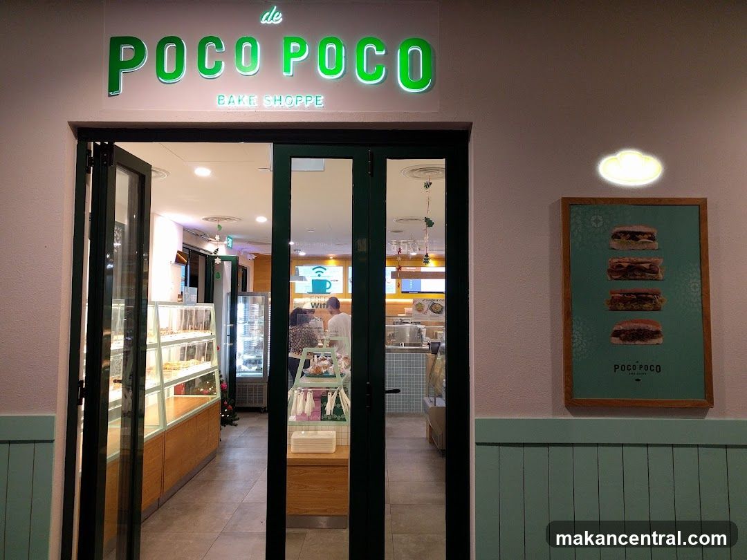 The exterior of a bake shoppe with a prominent green neon sign that reads "de POCO POCO BAKE SHOPPE". The entrance features double glass doors, with one set slightly ajar, revealing display cases filled with baked goods and a glimpse of the interior. To the right of the entrance, a framed poster displays four different sandwiches.