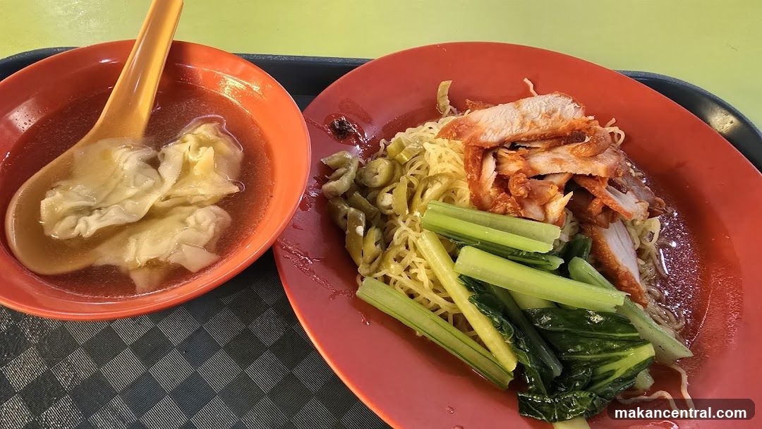 A close-up shot of a bowl of wonton noodle soup and a plate of char siu noodles. The soup contains wontons and is served with a bright orange spoon. The noodle plate features thin yellow noodles, slices of roasted pork (char siu), green vegetables, and pickled chilies. The food is presented on a black tray with a checkered pattern.