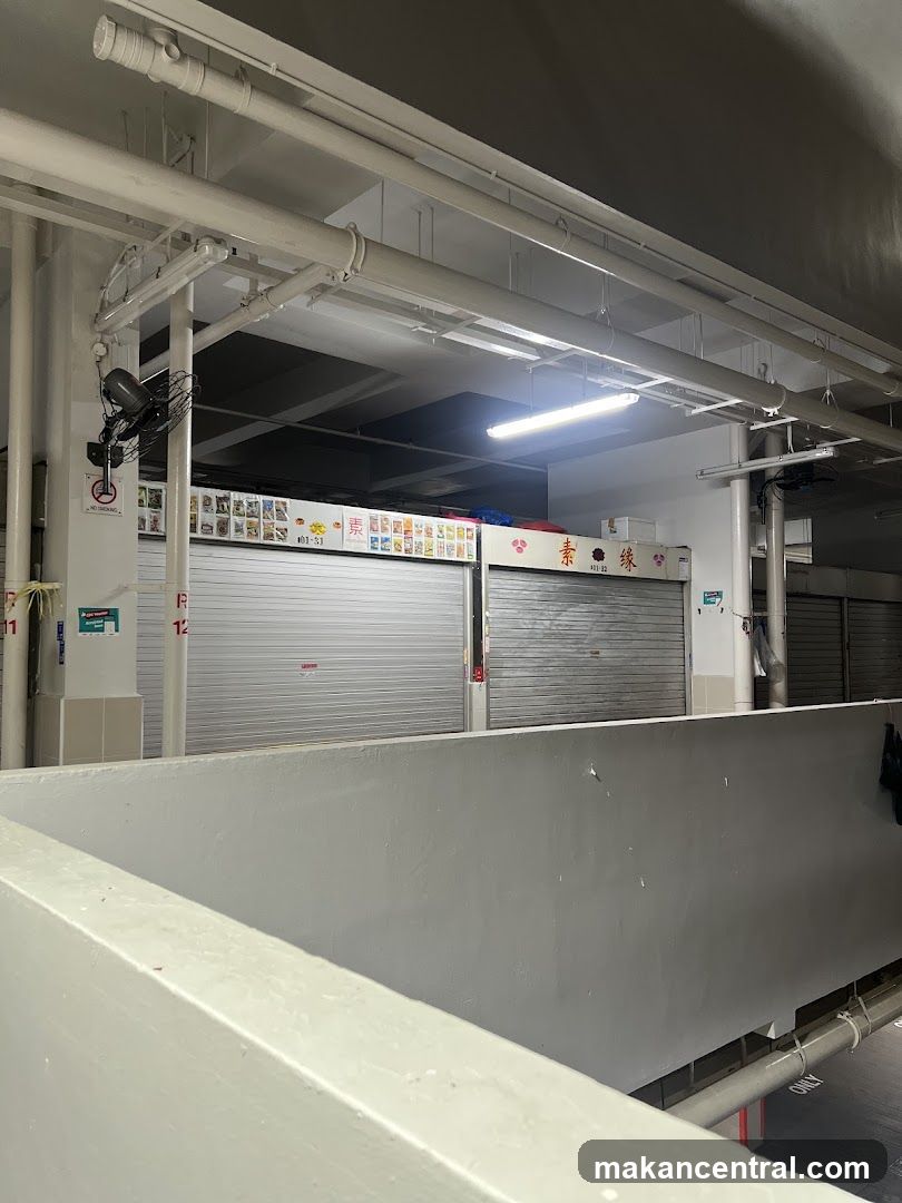 A row of closed market stalls with metal roll-down shutters. Above the shutters, there are signs with various images and text, some in Chinese characters. One stall has a sign with a red logo and Chinese characters that appear to read "素 缘". Another stall has the number "101-51" and a series of colorful images. A fan is mounted on a pillar to the left, with a "No Smoking" sign below it. The area appears to be an indoor market or food court.