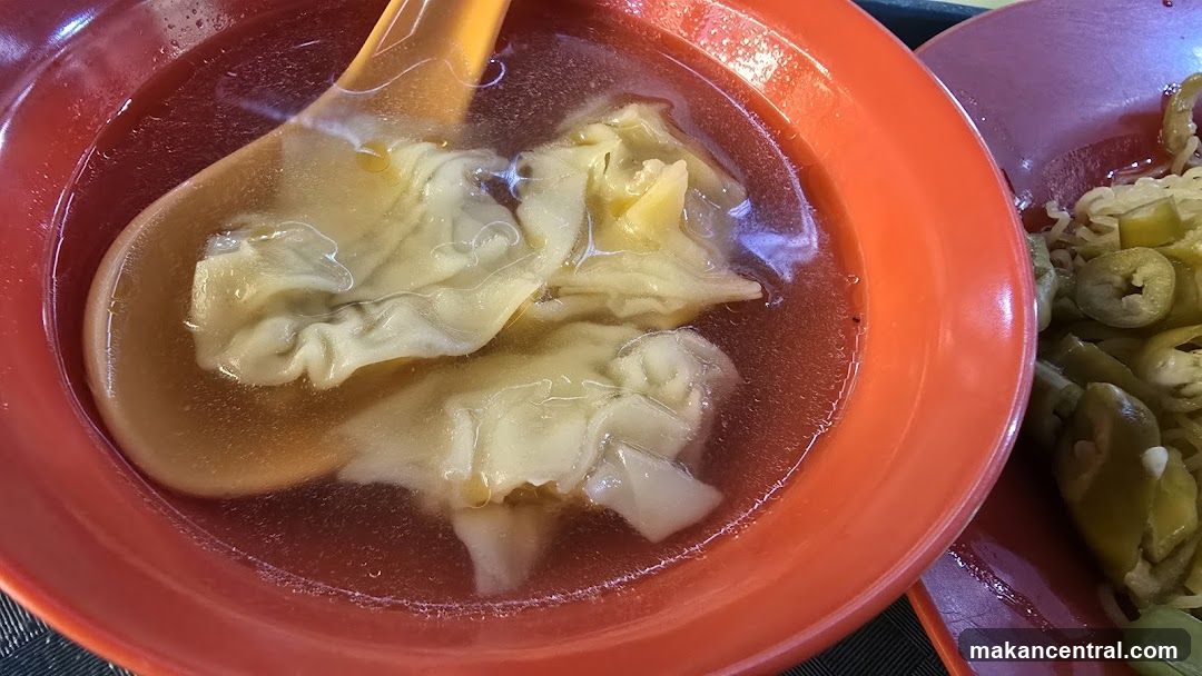 A close-up shot of wonton soup in a red bowl. A yellow spoon is partially submerged in the clear broth, holding several wontons. To the right, another red bowl contains noodles and sliced green peppers.