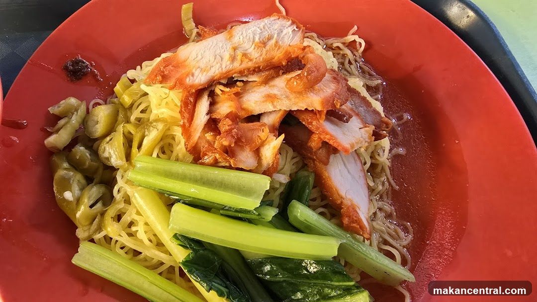 A close-up shot of a plate of char siu noodles. The dish features thin, yellow noodles in a savory broth, topped with slices of roasted char siu pork. Accompanying the noodles are blanched green vegetables and pickled green chilies. The food is served on a bright red plate.