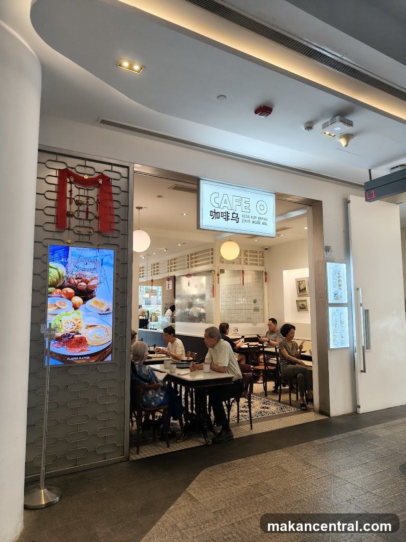 A modern cafe entrance with a white sign displaying 'CAFE O' in English and Chinese characters above the doorway. The interior is visible, showing patrons seated at tables, warm lighting, and traditional decor elements like red tassels on a patterned wall panel. A digital display to the left shows food images under 'ALL DAY SPECIAL'. The floor has a tiled pattern, and ceiling lights are recessed.