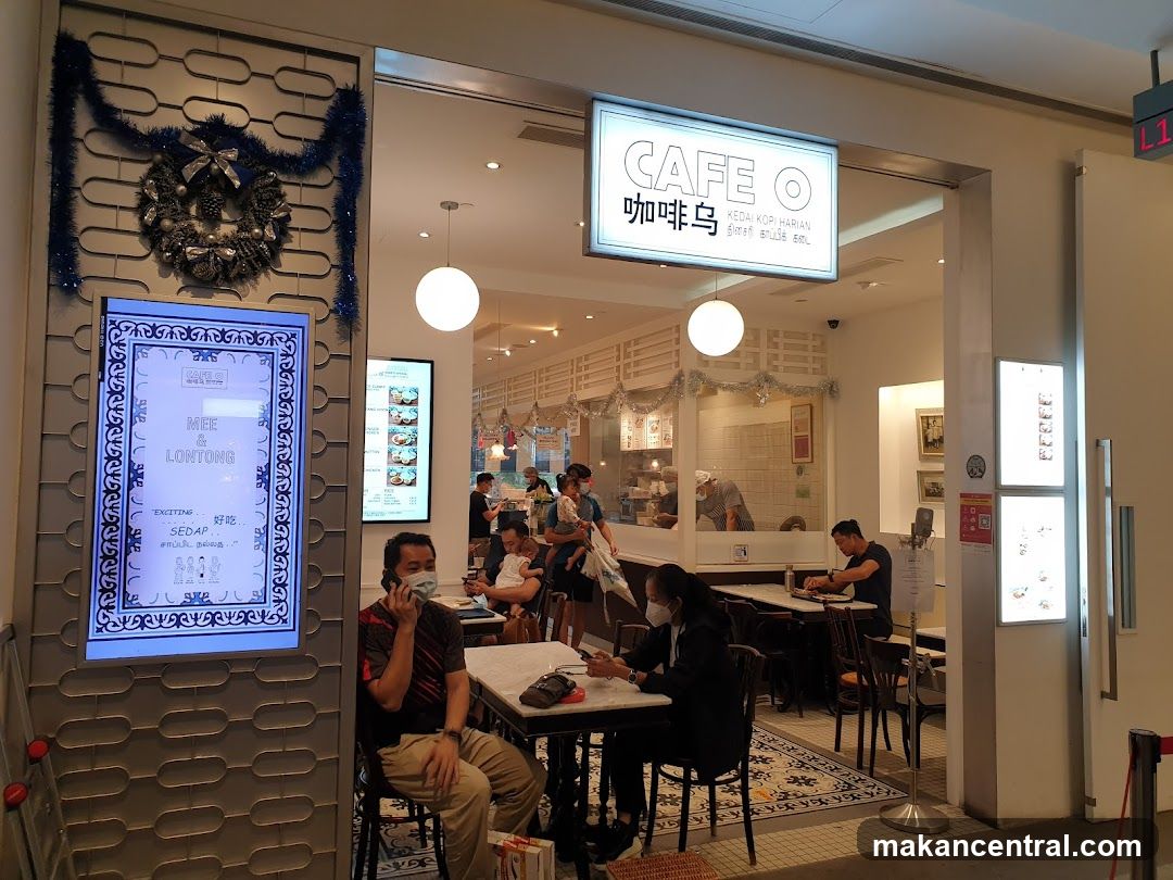 Exterior view of a cafe entrance with illuminated sign reading 'CAFE O' and Chinese characters '咖啡鸟'. The facade features decorative blue tinsel and a wreath above the door. Inside, customers are seated at tables, some wearing masks. A digital menu board on the left displays 'MEE & LONTONG' and 'EXCITING SEDAP'. The interior has bright lighting, white walls, and patrons dining near a counter.