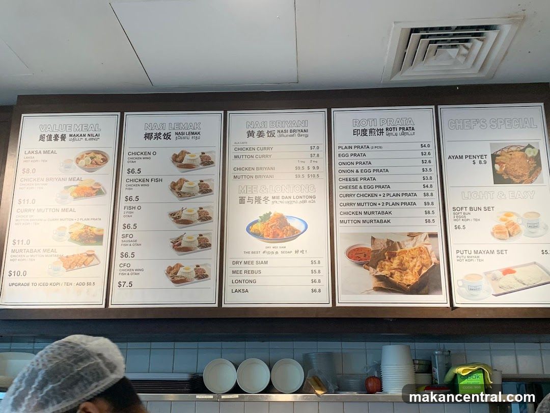 A wall-mounted menu board displaying various food categories in English and Chinese with prices. Sections include Value Meal, Nasi Lemak, Nasi Briyani, Roti Prata, Chef's Special, and Light & Easy. Below the menu, a kitchen counter holds stacked plates, cups, and a green branded container.