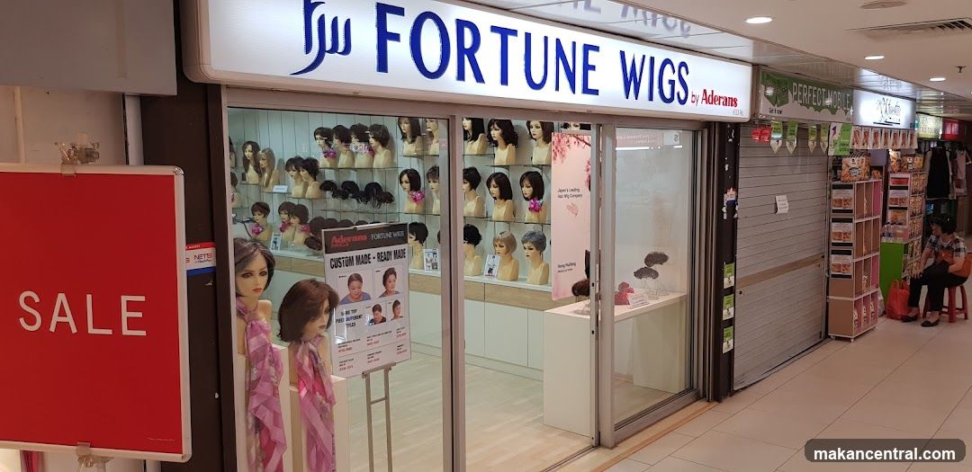 Storefront with illuminated sign reading 'FORTUNE WIGS by Aderans'. Glass display windows show mannequin heads with wigs. A red sale sign is on the left. Adjacent store has a shuttered entrance.
