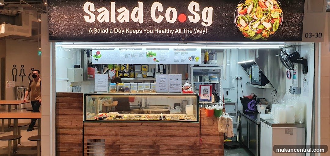 A salad shop with a wooden counter and glass display case filled with salads. The sign above reads 'Salad Co.Sg' and includes the slogan 'A Salad a Day Keeps You Healthy All The Way!'. A photo of a colorful salad is displayed on the right side of the sign. Behind the counter, there are shelves with jars, a menu board, and kitchen equipment. A person wearing a face mask walks past to the left.