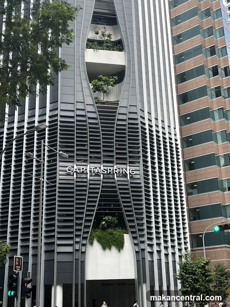 Modern high-rise building facade with vertical slatted design and integrated greenery. The name 'CAPITASPRING' is prominently displayed in white lettering on the exterior. Below it, 'Hawker Centre Levels' is visible above an entrance. Adjacent buildings and street elements like traffic lights and trees are present.