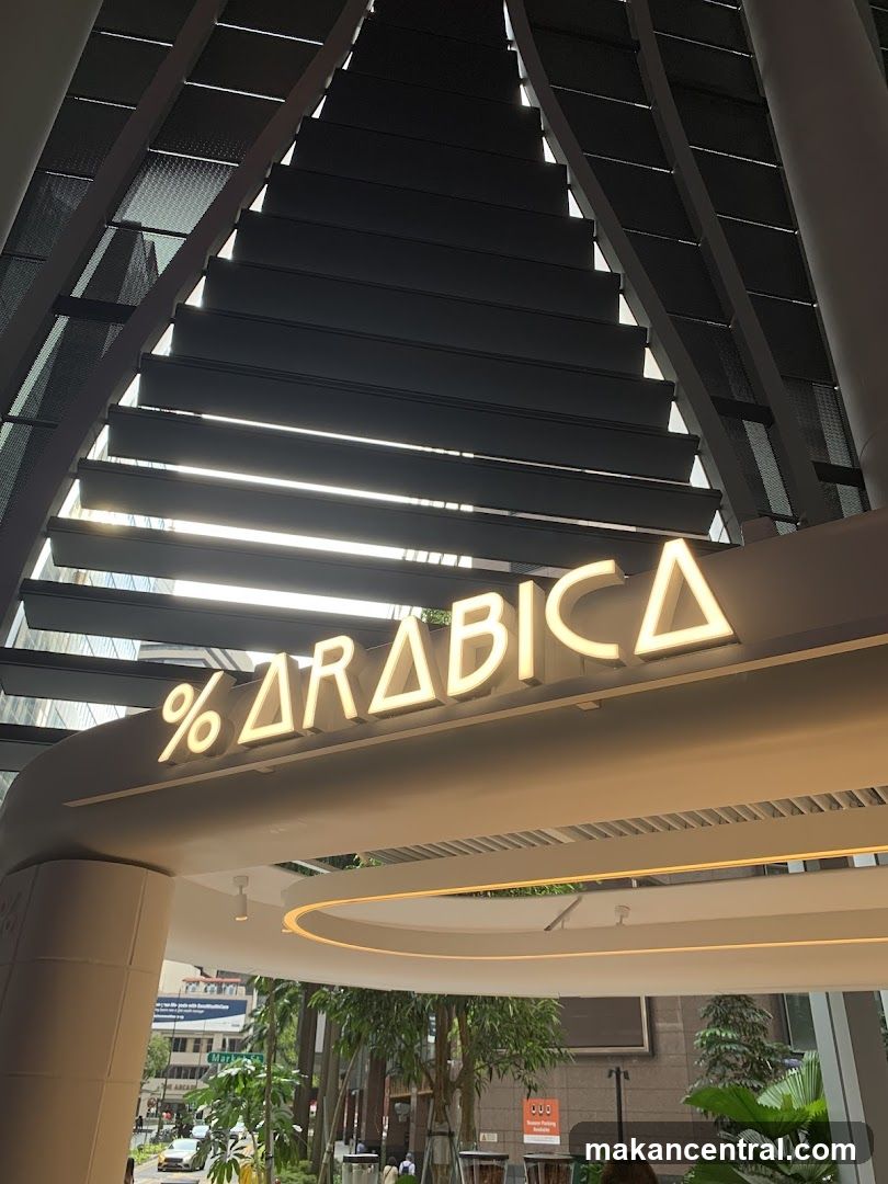 Modern architectural entrance with illuminated signage featuring the logo '% ARABICA' in glowing yellow letters. Above is a slatted metal canopy structure allowing light to filter through. Visible greenery and urban street elements beyond the entrance.