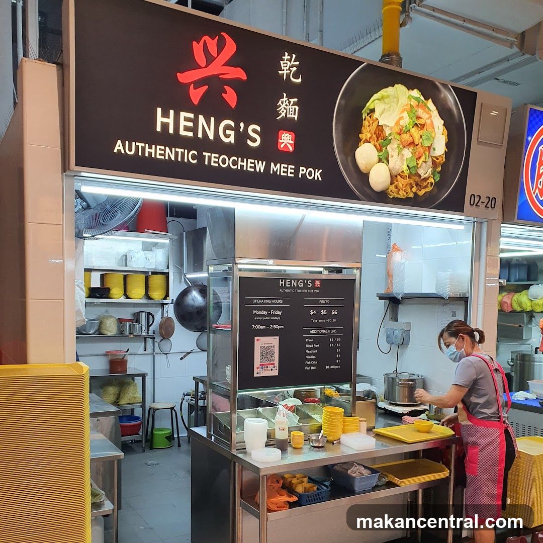 A food stall with a black sign featuring red Chinese characters '兴乾麵' and the English name 'HENG'S AUTHENTIC TEOCHEW MEE POK'. A large photo of a noodle dish is displayed on the right side of the sign. The stall has a stainless steel counter, yellow bowls stacked nearby, and an open kitchen area behind glass. A staff member wearing a face mask and apron is preparing food at the counter.