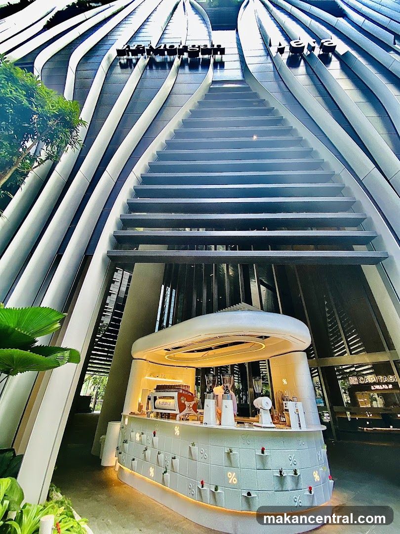 Low-angle shot of a contemporary building facade featuring curved metallic slats and a central staircase leading upward. Below, a circular white coffee counter with embedded % symbols and small plant holders is illuminated. Greenery frames the left side. Signage reading 'MORGAN' and 'AERI' is mounted on the upper structure.