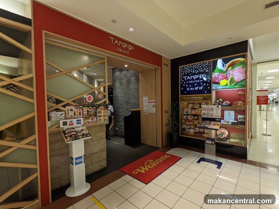 The exterior entrance of Tampopo Grand restaurant. The entrance features a red frame with wooden accents. Above the entrance, the restaurant's name "Tampopo Grand" is displayed. To the right of the entrance, there is a display showcasing various food items and promotions. A red welcome mat is placed at the entrance.