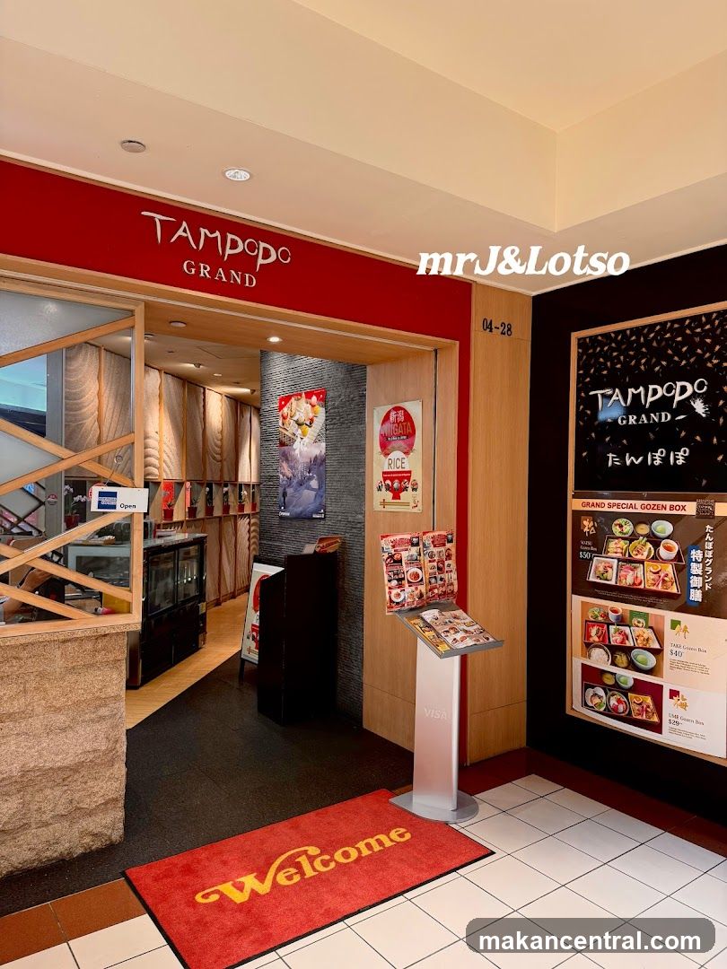 The entrance to Tampopo Grand restaurant features a red facade with the restaurant's name in white lettering. A welcome mat is placed on the floor. To the right, a menu board displays various gozen box options with prices. Inside, a display case and decorative walls are visible.
