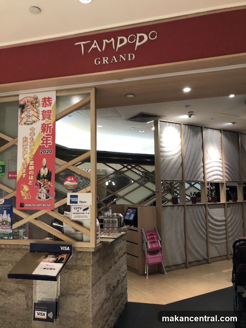 The exterior of a restaurant called Tampopo Grand. A red sign with the restaurant name is visible above the entrance. To the left of the entrance, a poster with Japanese text and the year 2020 is displayed. A stand with Visa payment information is also present.